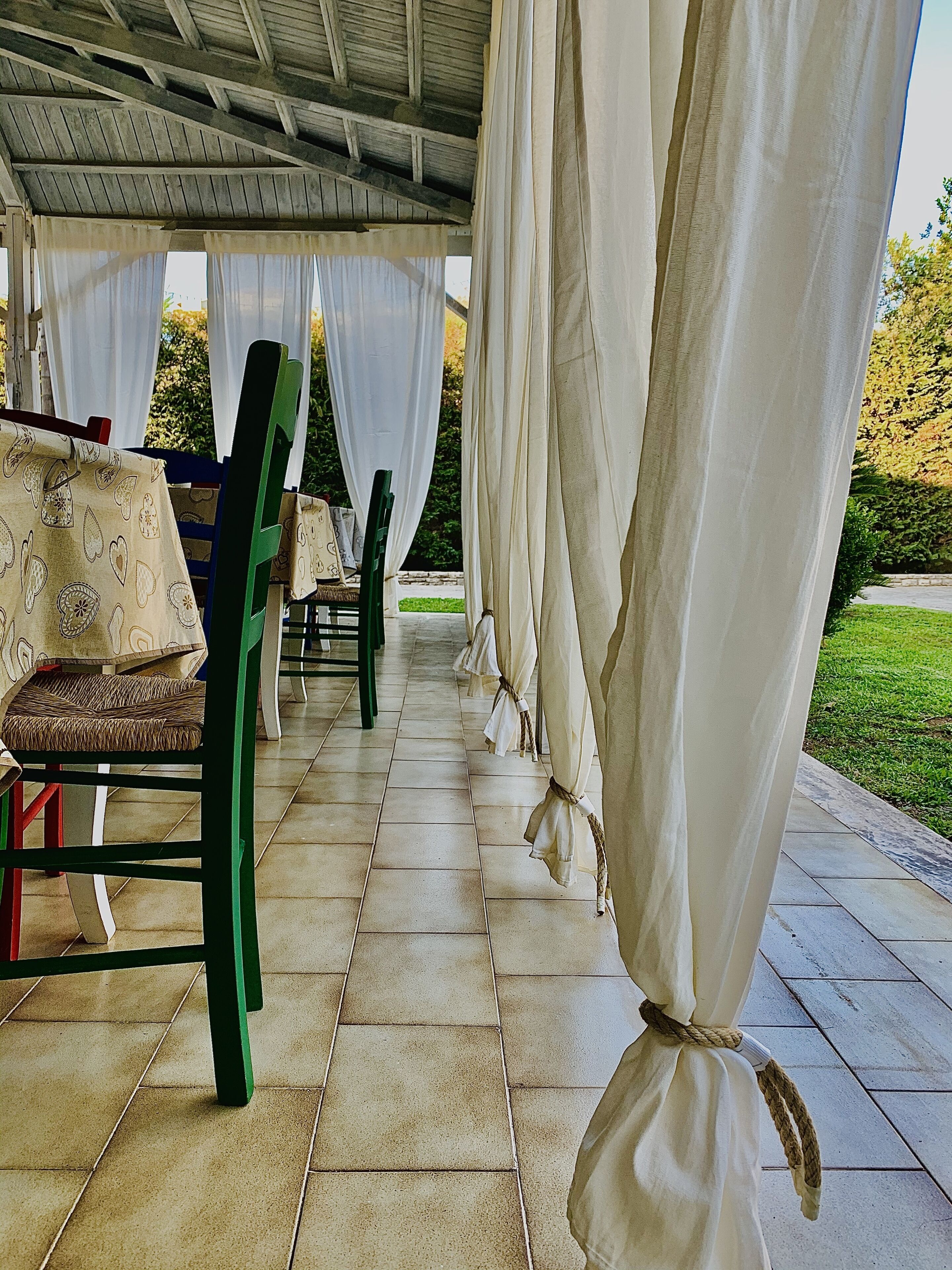 Un espace extérieur aménagé avec des sièges, des chaises et des tables, des rideaux blancs et un sol carrelé.