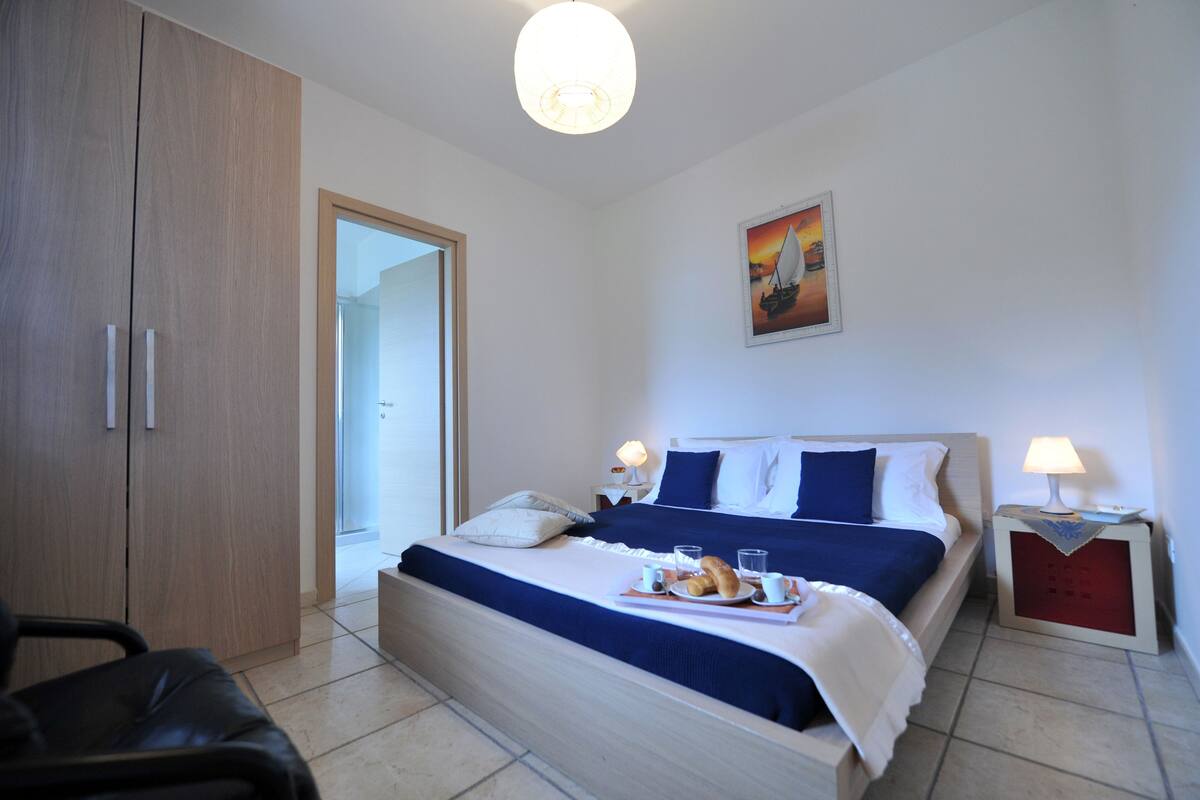A hotel room with a bed, bedside tables, a chair, a wardrobe, and a painting on the wall.