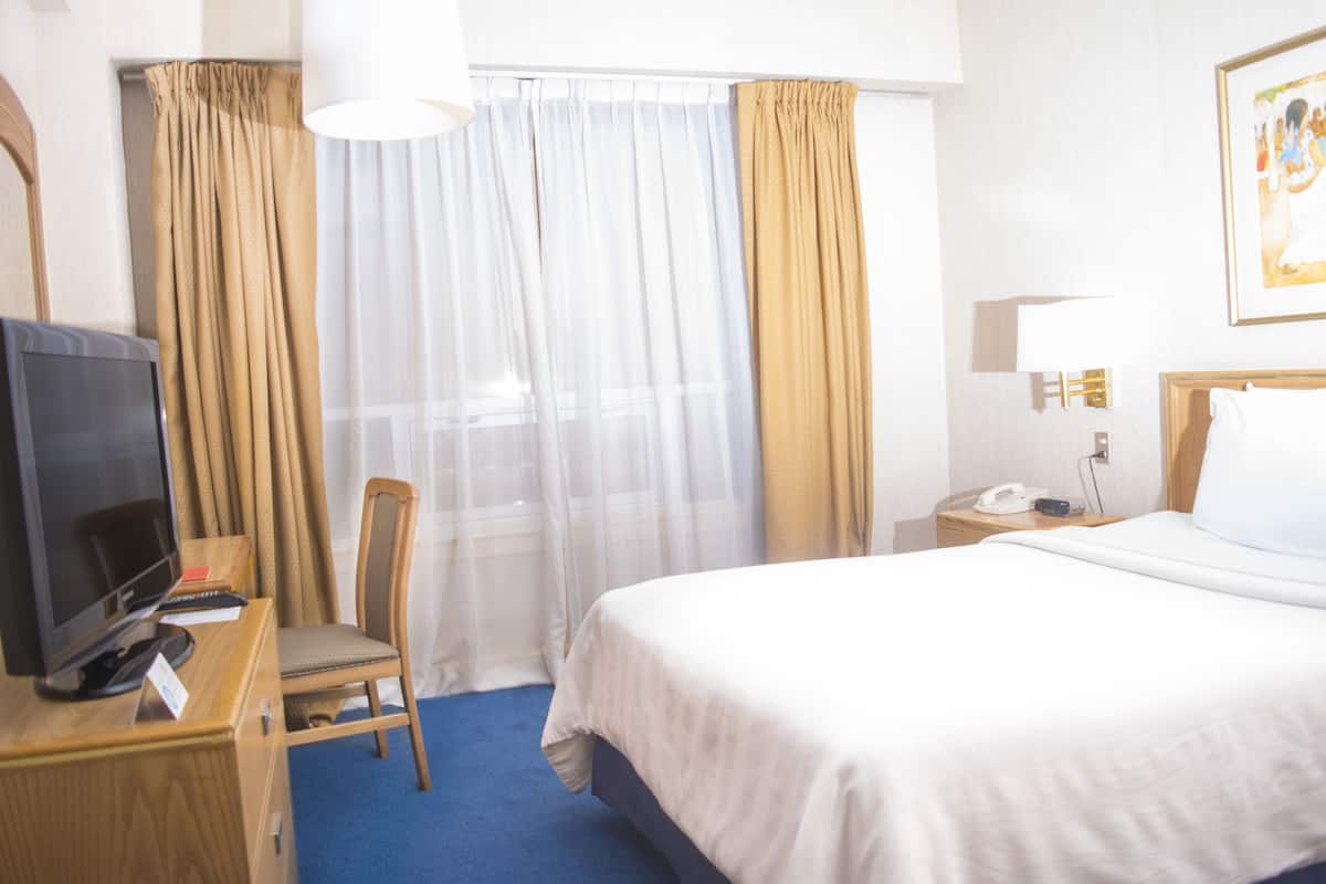 Una habitación de hotel con cama, escritorio con televisor, silla, mesita de noche con teléfono y ventana con cortinas.