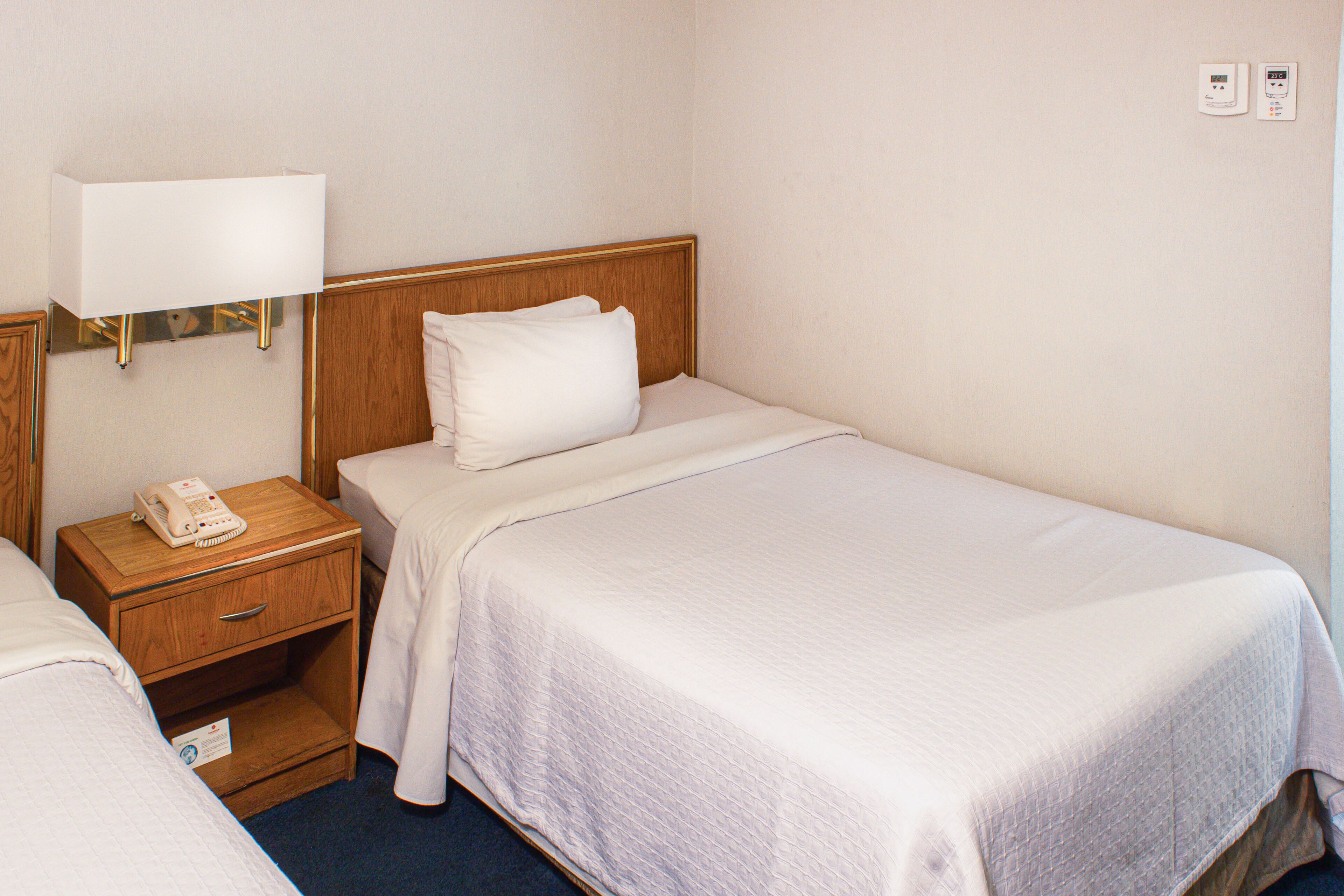 A hotel room with two single beds, a wooden nightstand with a telephone, a wall-mounted lamp, and a thermostat on the wall.