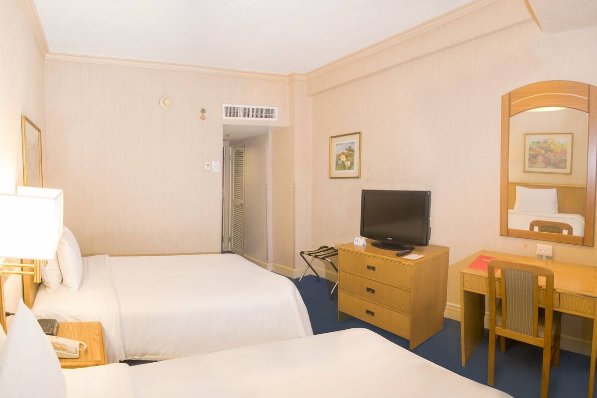 Habitación de hotel con dos camas, un escritorio, televisión, espejo y una puerta que da a otra habitación.