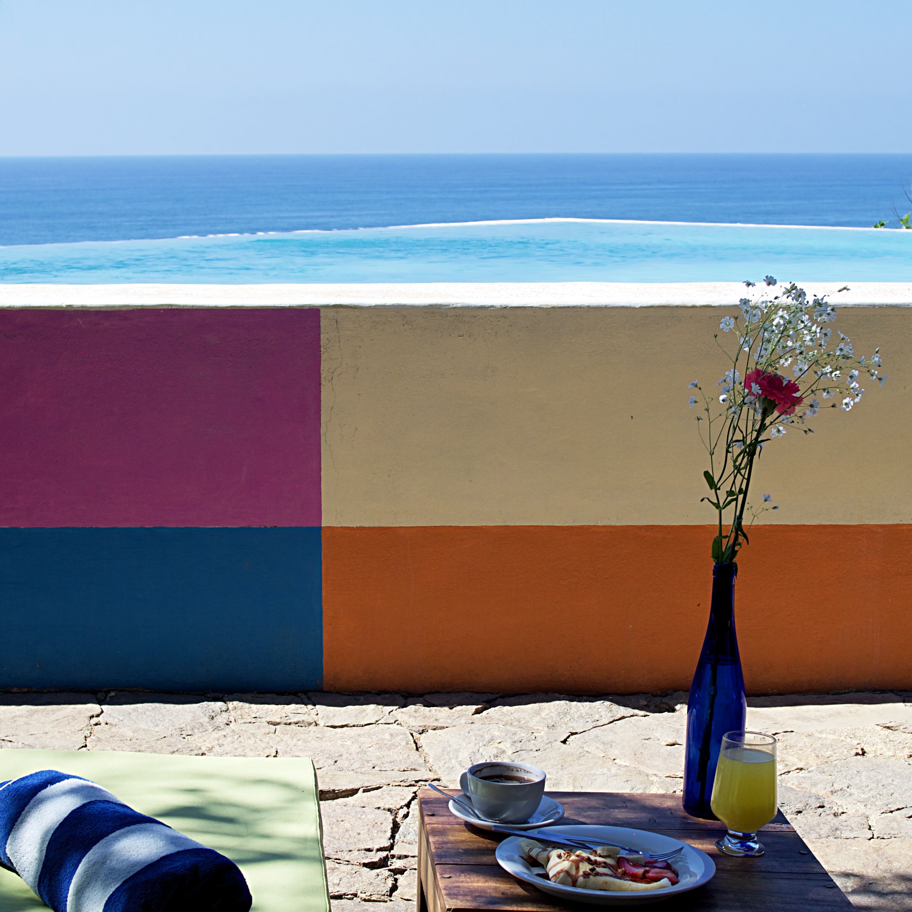 A table with a cup, saucer, plate with food, and a glass of juice, set against a colorful wall with a view of the ocean.