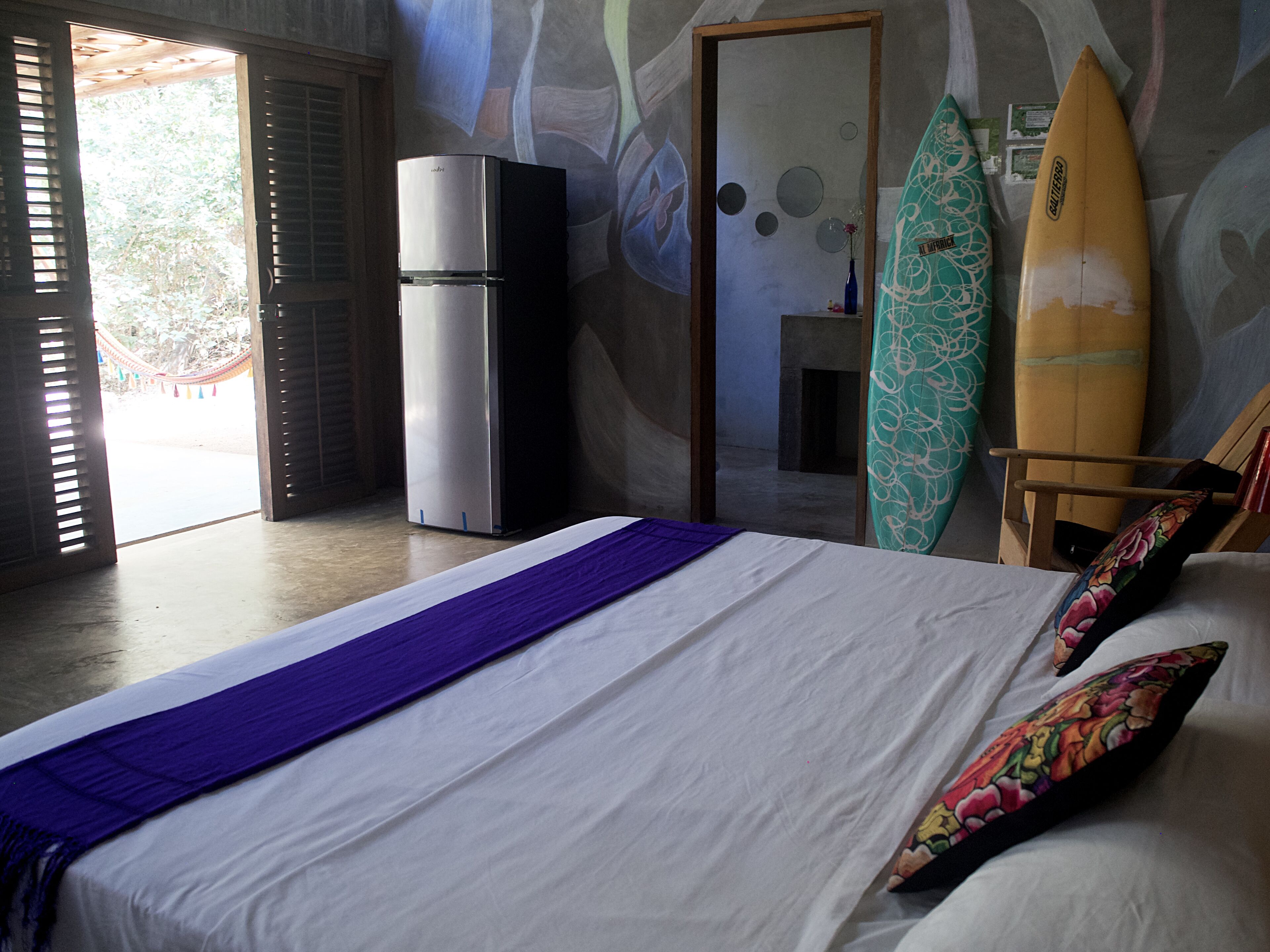 A hotel room with a bed, a refrigerator, a surfboard, and a view of the outdoors.