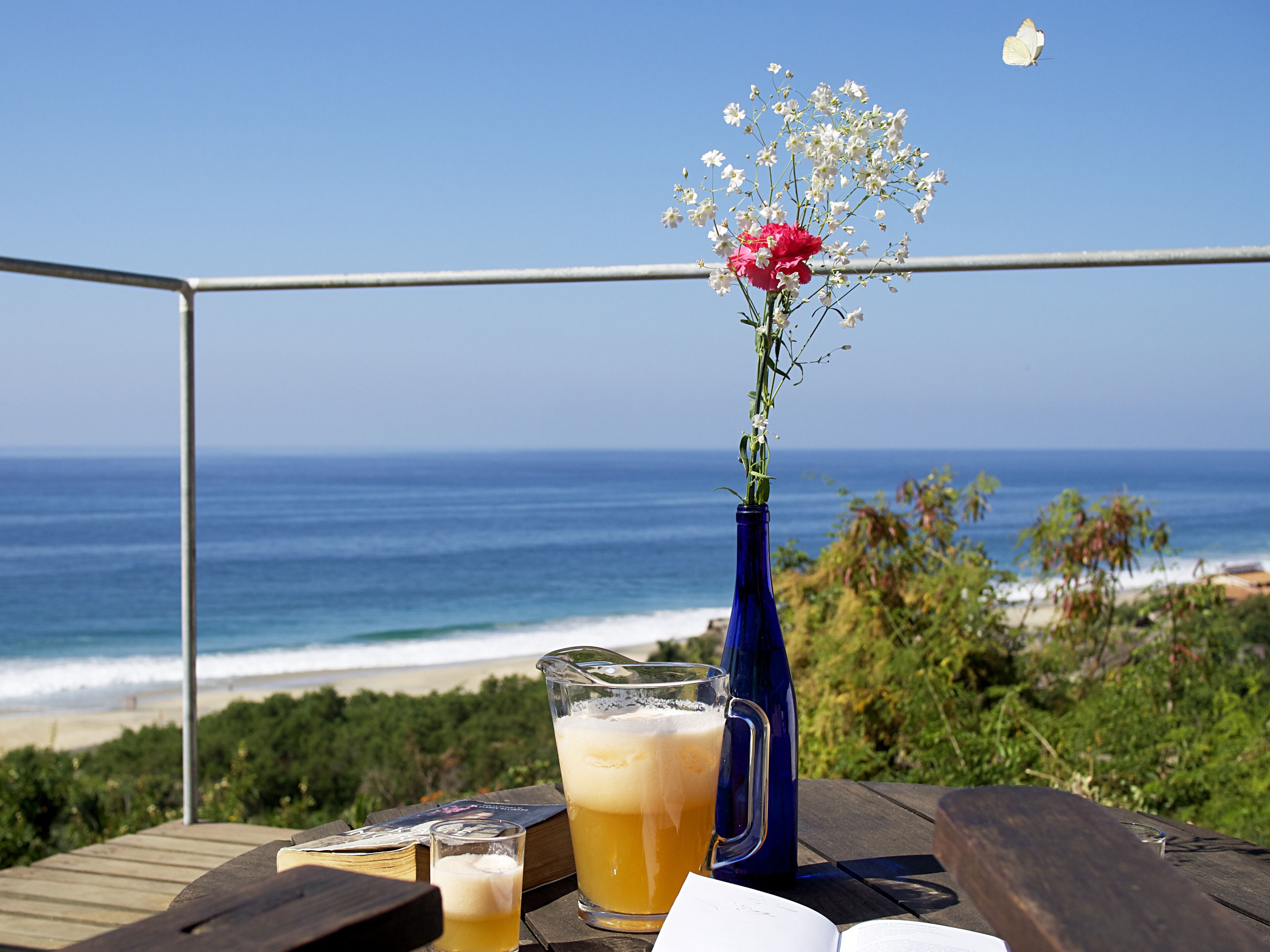 A table with drinks and a vase of flowers, overlooking a beach.