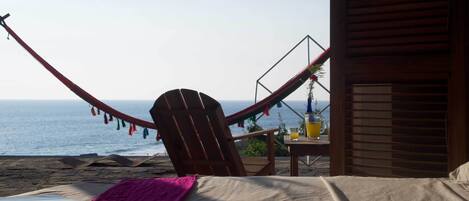 A hammock chair facing the ocean, a bed with a pink blanket, and a table with a pitcher of juice.