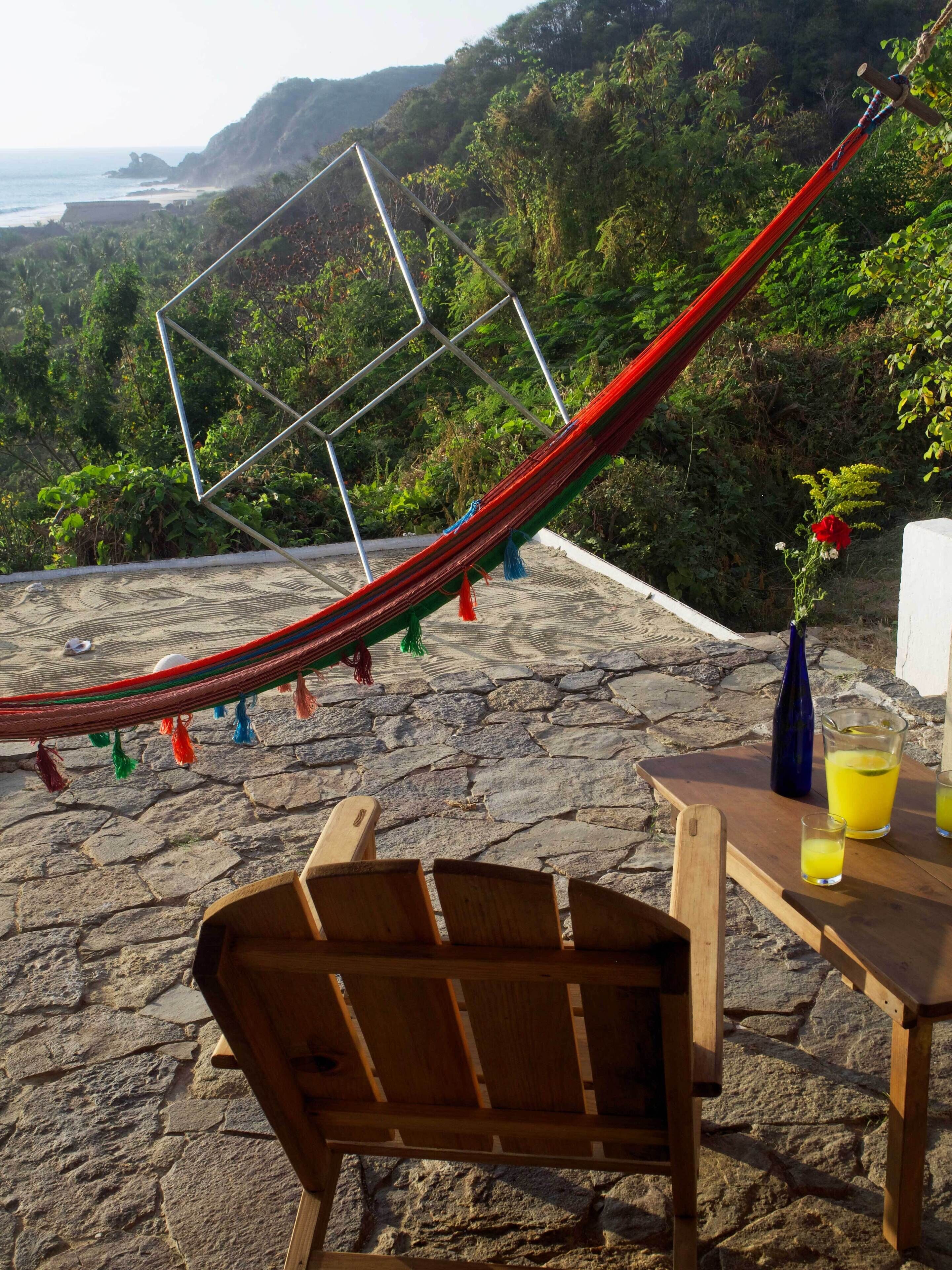 A hammock, wooden chairs, and a table with drinks on a stone patio overlooking a beach and forested area.