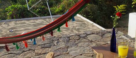 A hammock, wooden chairs, and a table with drinks on a stone patio overlooking a beach and forested area.