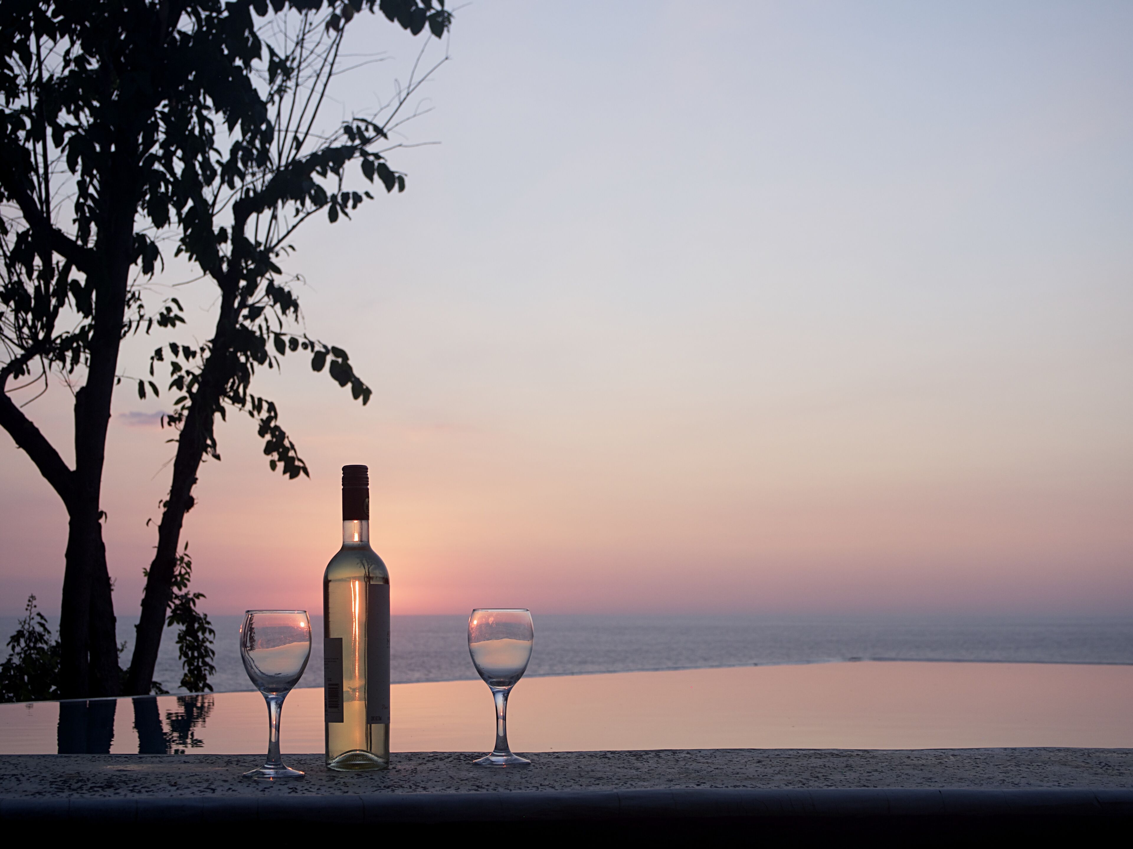 A bottle of wine and two glasses on a table with a view of the ocean at sunset.