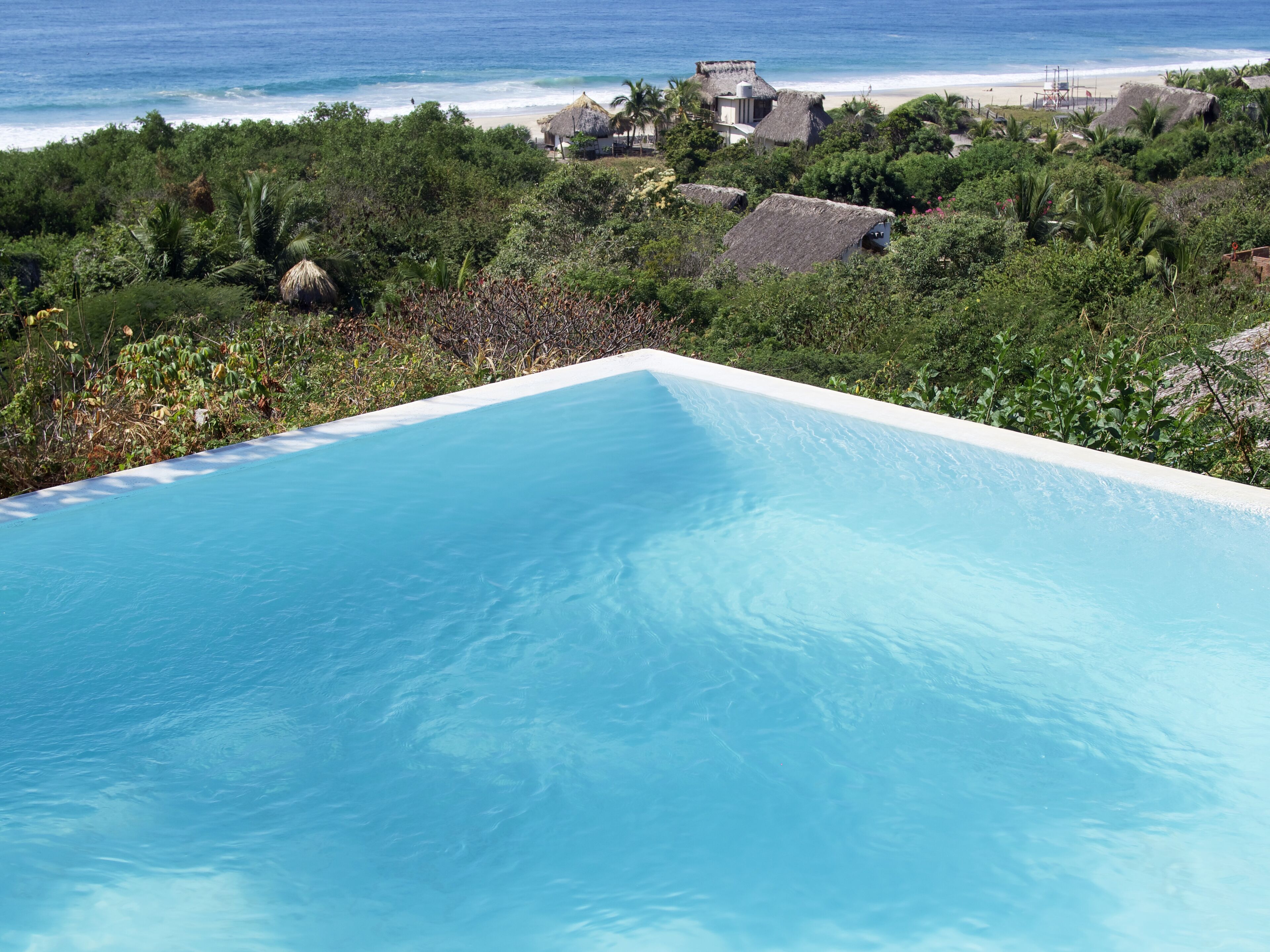 Une piscine d’un bleu éclatant, avec vue sur l’océan et les maisons de plage en arrière-plan.