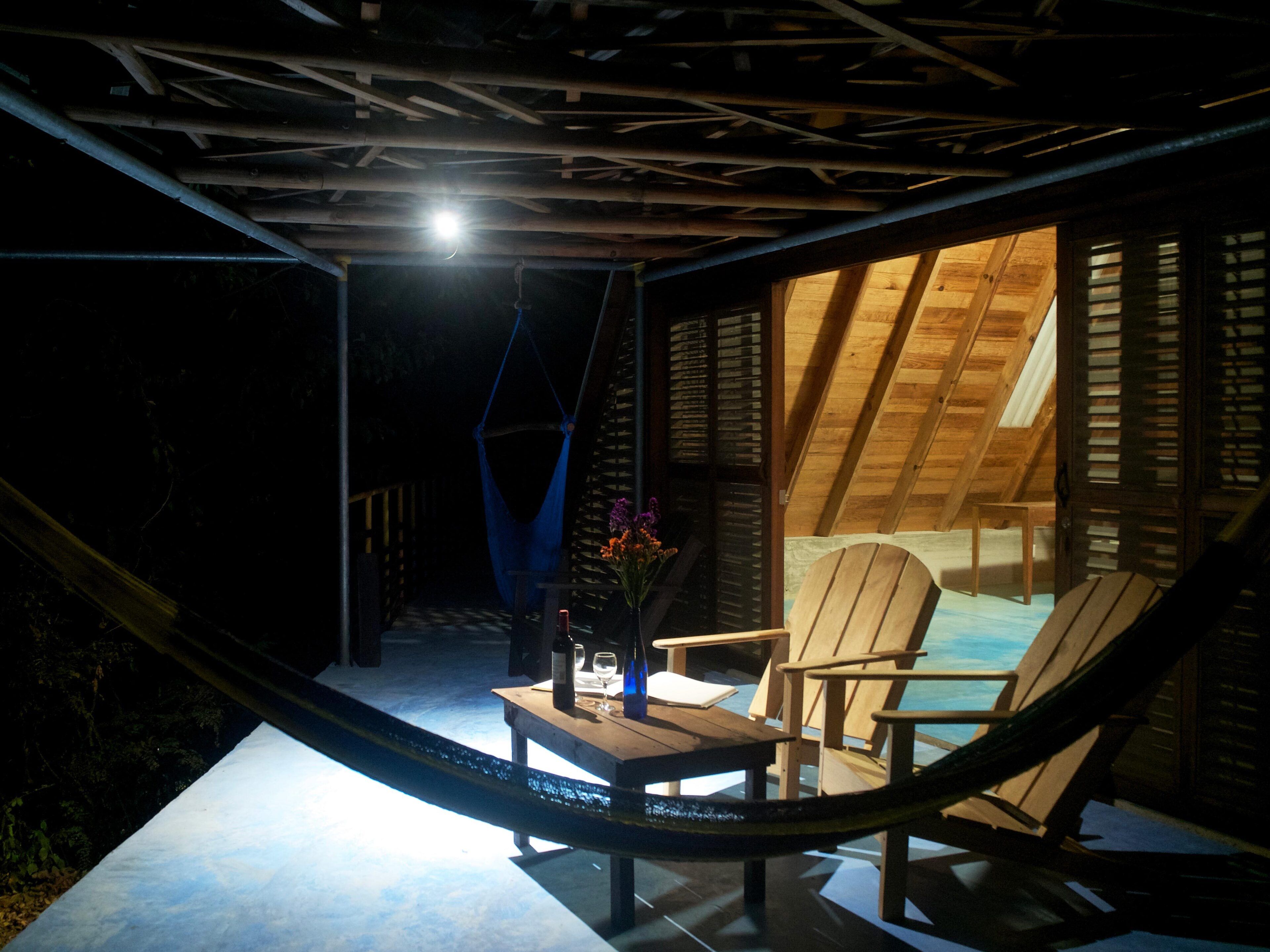 A hammock chair and a wooden table with a bottle and glass, under a sloped roof with wooden beams.