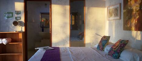 A bedroom with a bed, a wooden shelf, a mirror, a framed picture, and a window.