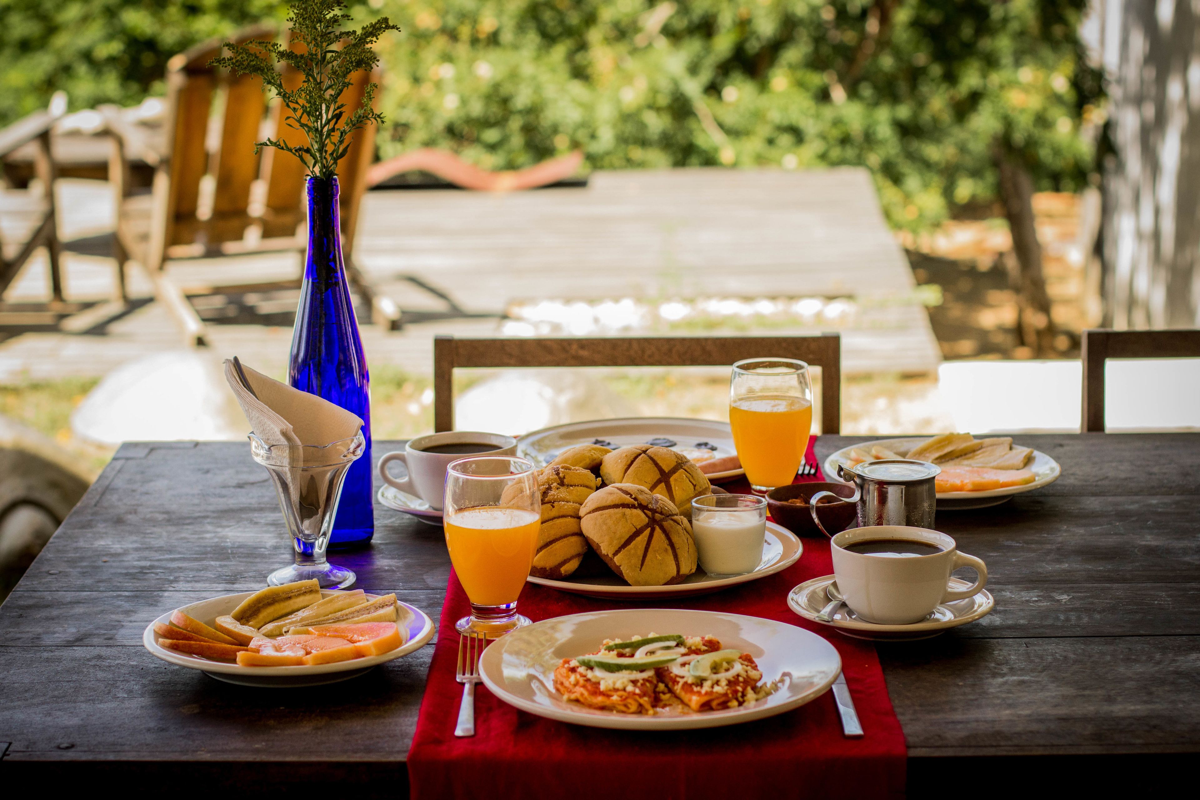 Una mesa de madera dispuesta con cosas para el desayuno, como pan, pasteles y un plato de comida, junto con un jarrón azul y una taza de café.