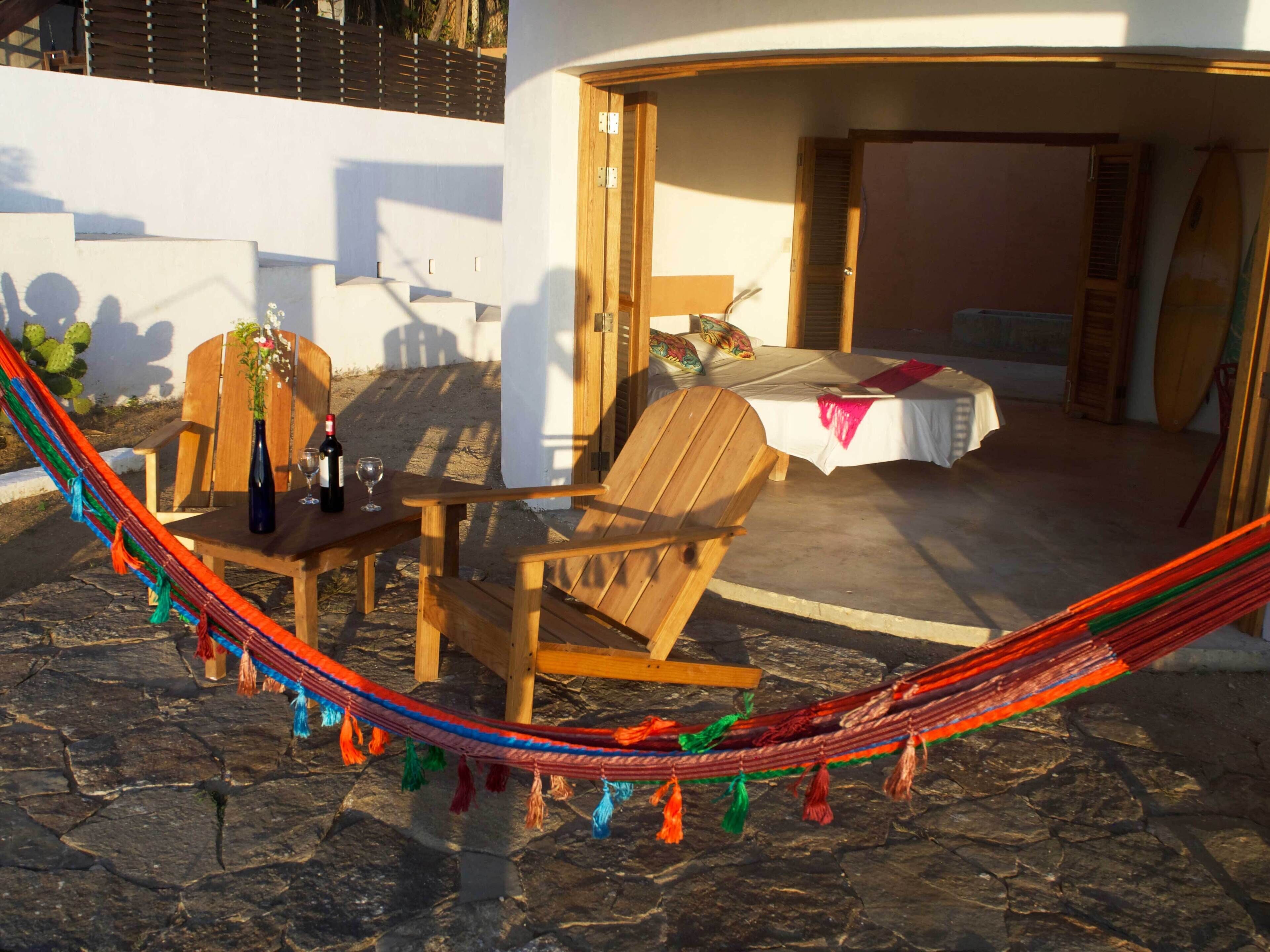 A hammock between two wooden chairs and a small table with a bottle and glass, set against a white wall with a bed in the background.