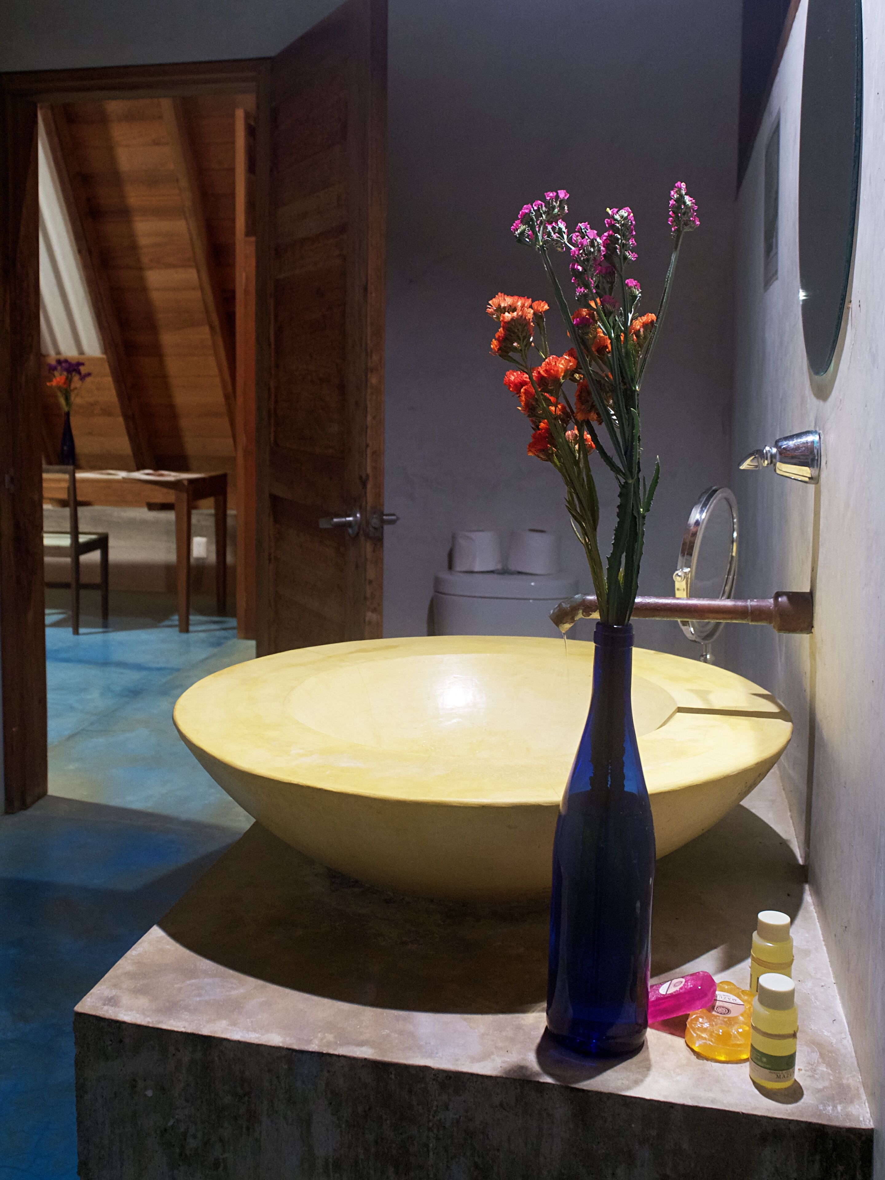 A large yellow sink with a blue vase containing flowers, a mirror, and toiletries on a countertop.