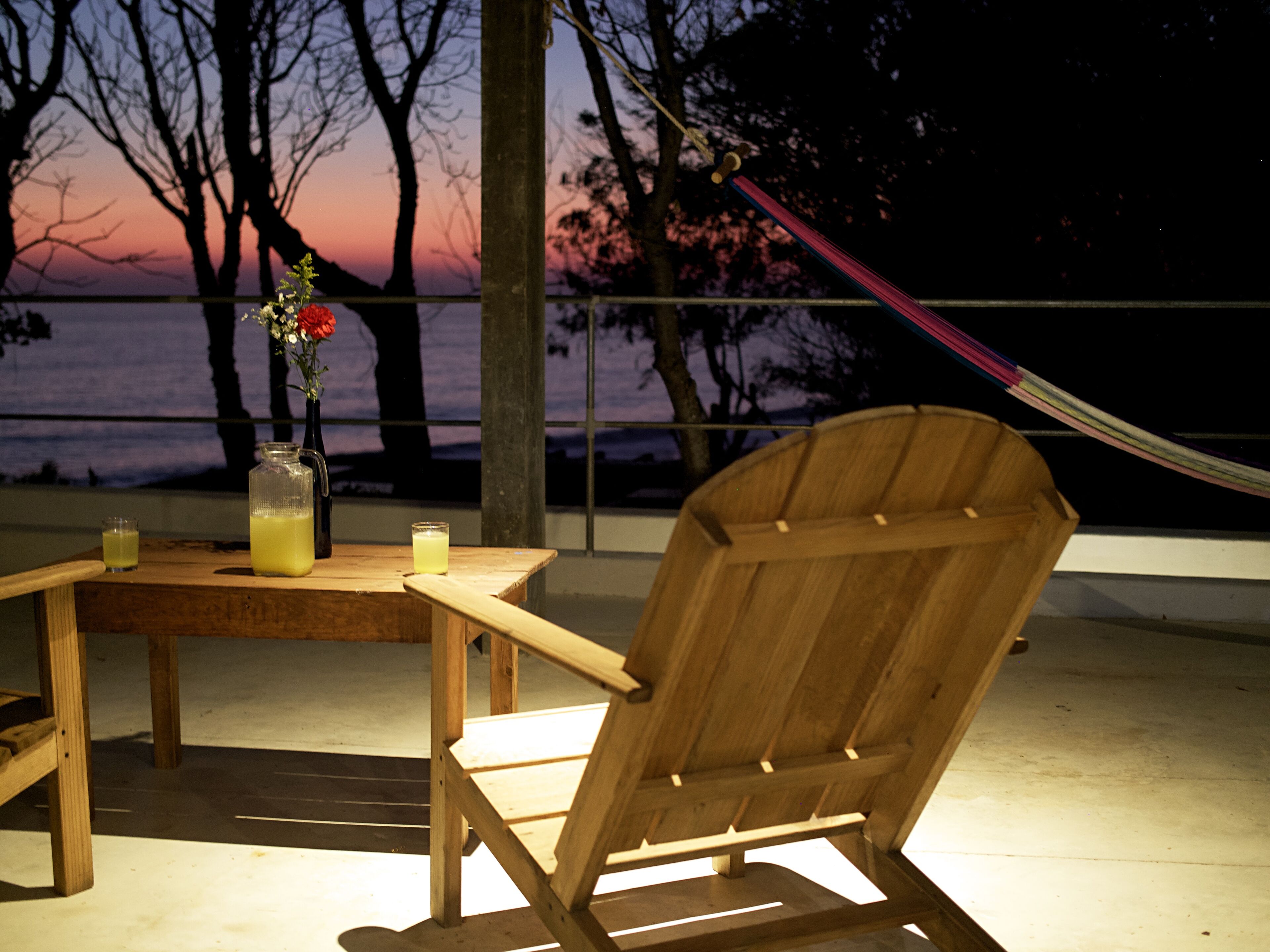 A wooden chair and table on a balcony with a view of trees and a sunset.
