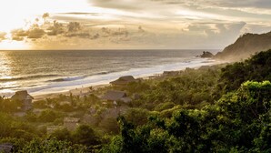 Un paisaje costero con una playa, cabañas de techo de paja y abundante vegetación.