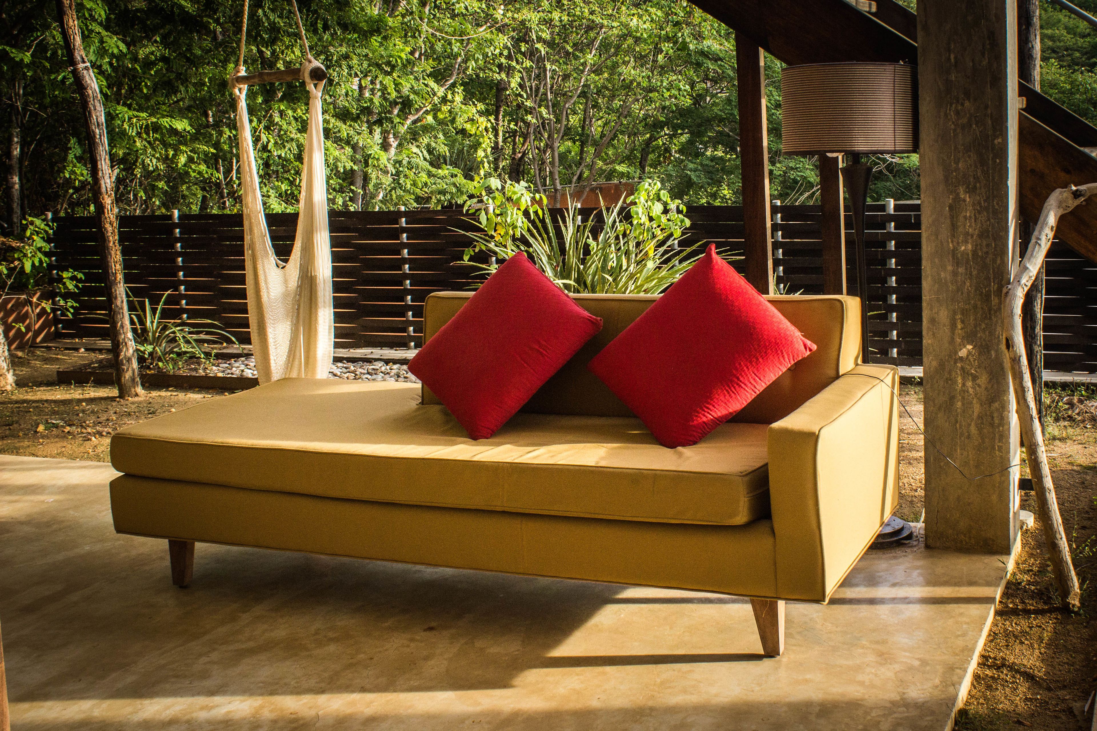 A yellow sofa with red cushions in an outdoor setting with a wooden deck and a metal railing.