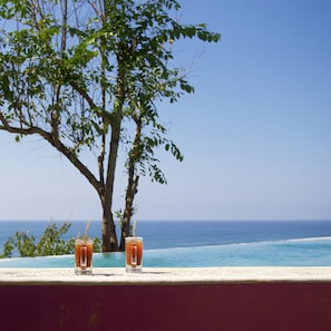 Two glasses of orange beverage on a ledge with a view of the ocean and a tree.