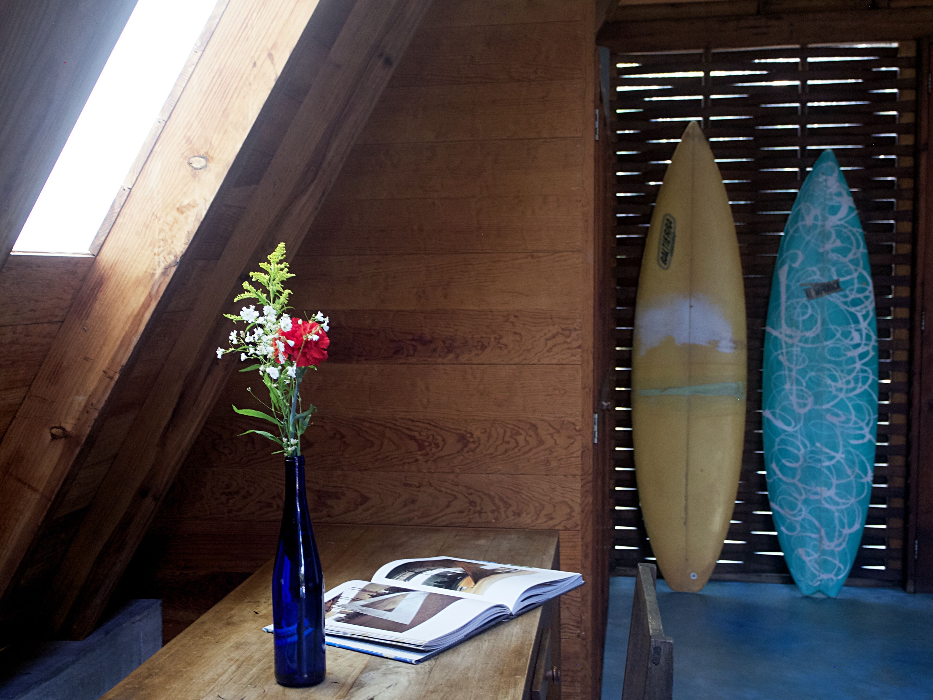Une table en bois, sur laquelle se trouvent un vase bleu rempli de fleurs, une planche de surf et un magazine ouvert.