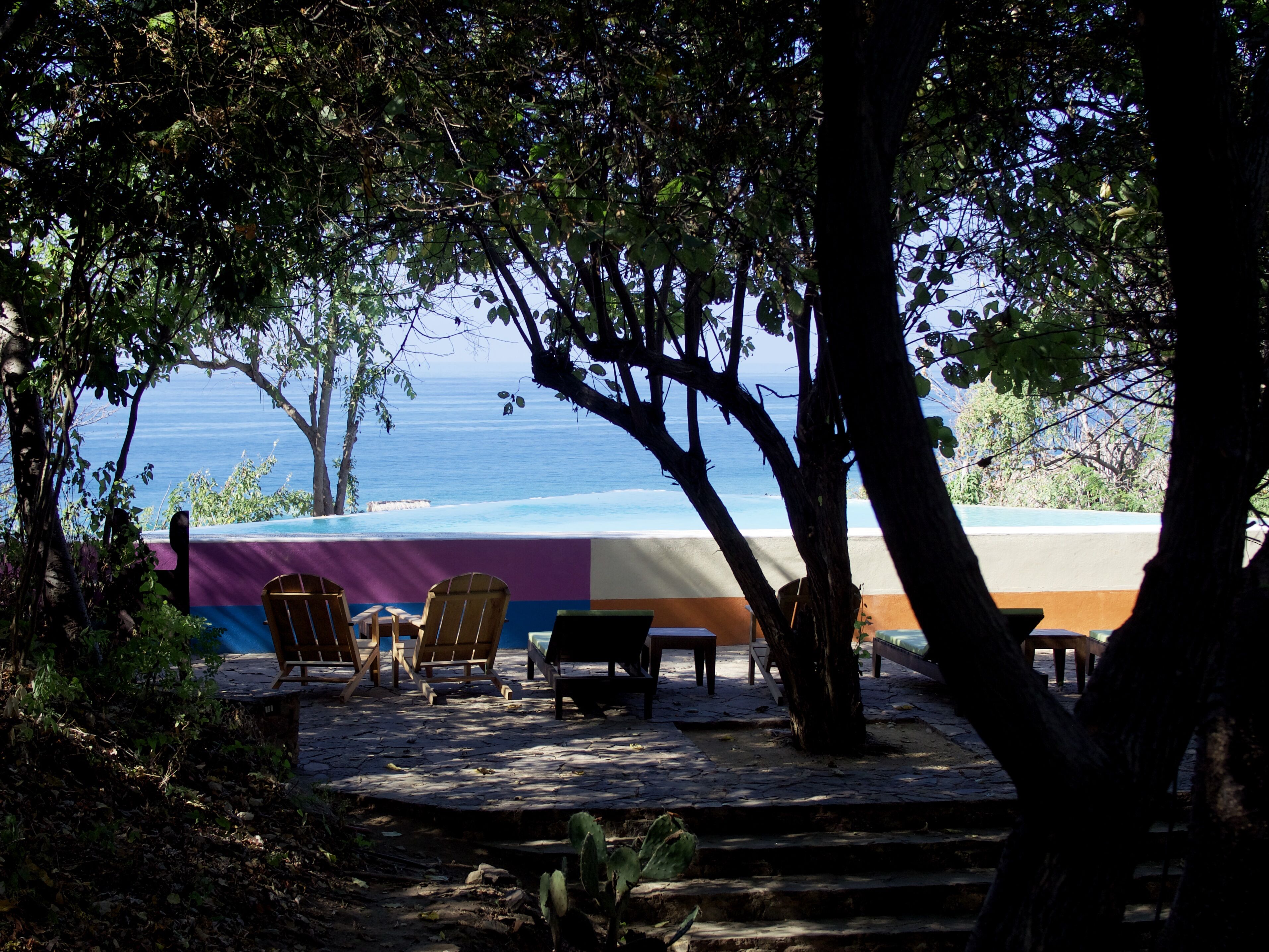 A patio with wooden chairs and a table, overlooking a pool and the ocean.