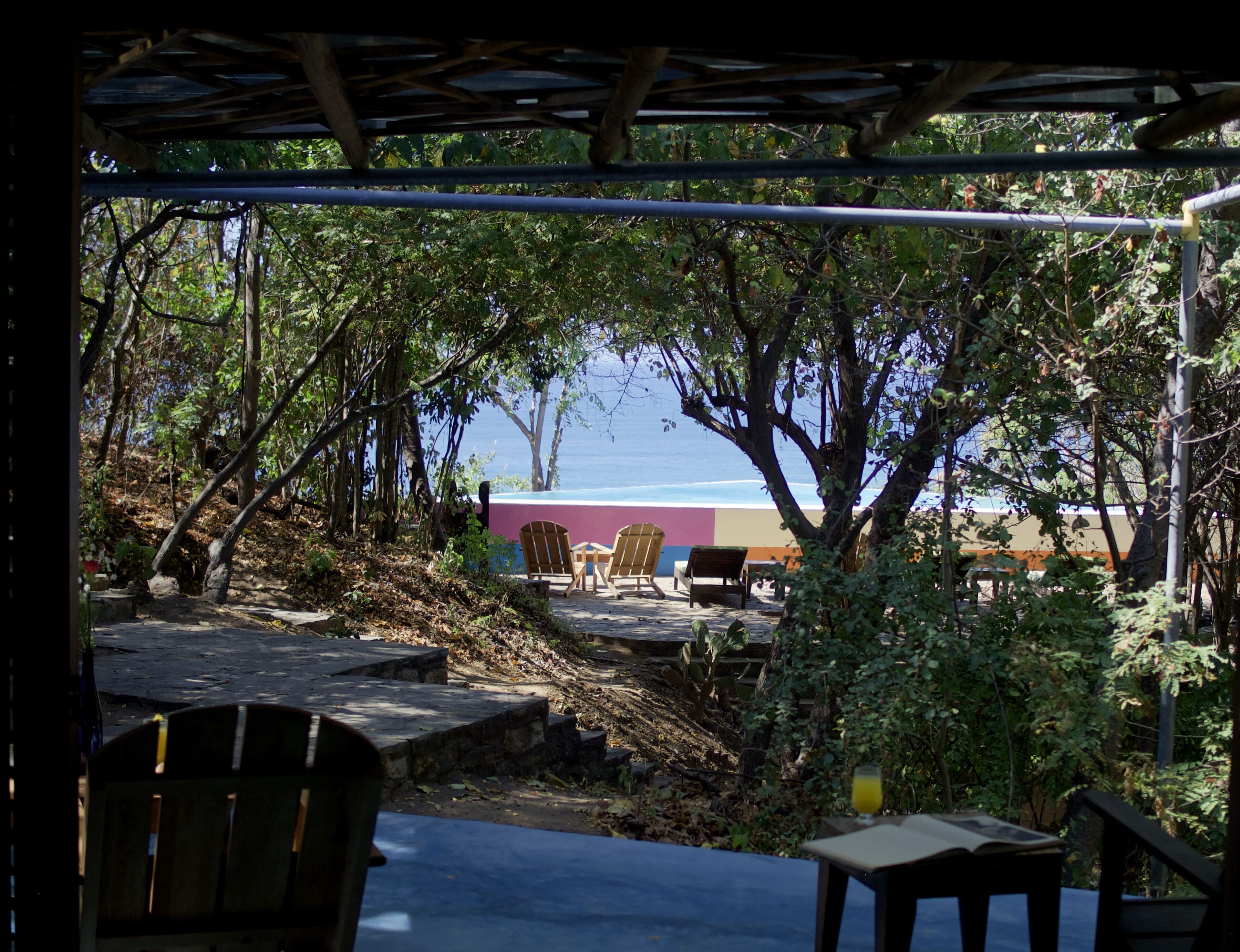 A covered outdoor seating area with wooden chairs and tables, surrounded by trees and overlooking a beach.