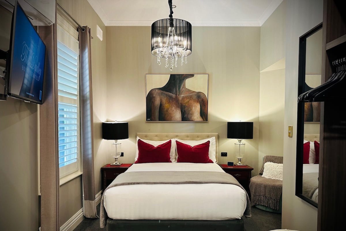 A hotel room with a large bed, two bedside tables with lamps, a mounted TV, a chandelier, a painting on the wall, and a mirror.