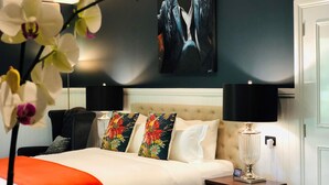 Una habitación de hotel con cama, mesita de noche, lámpara, un cuadro en la pared y una planta de orquídea.