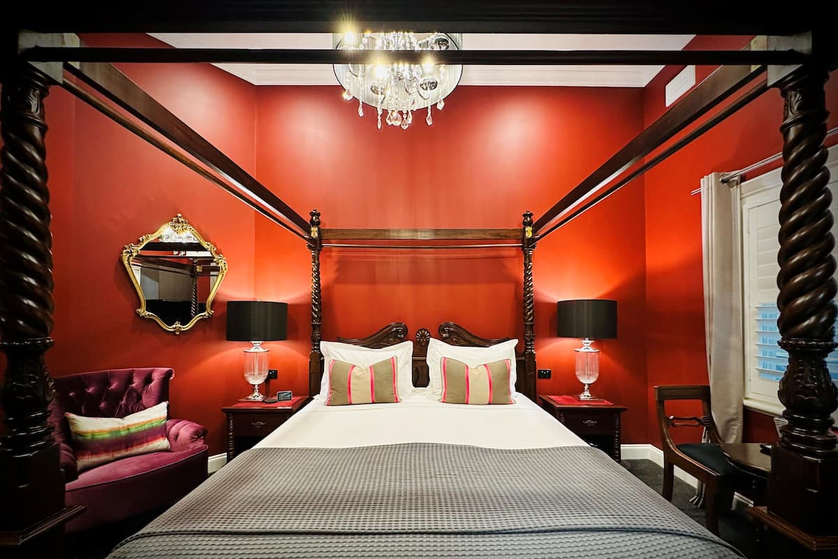 A four-poster bed with a canopy, a mirror, two bedside tables with lamps, a red upholstered chair, and a chandelier.