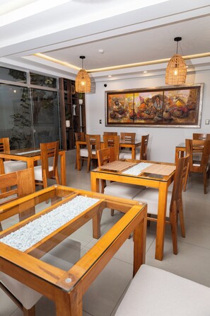 Un área de comedor con mesas y sillas de madera, una mesa con cubierta de vidrio y guijarros blancos, y un cuadro abstracto grande en la pared.