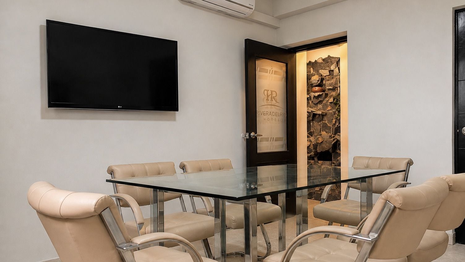 Una sala conferenze con un tavolo in vetro e poltrone in pelle, una TV a schermo piatto montata a parete e un muro in pietra con una porta che reca la scritta 'RIVERA DEL RIO HOTEL'.