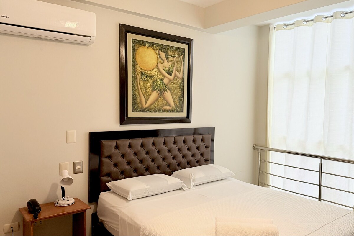 Una habitación de hotel con cama, mesita de noche, unidad de aire acondicionado, una obra de arte enmarcada y una ventana con cortinas.