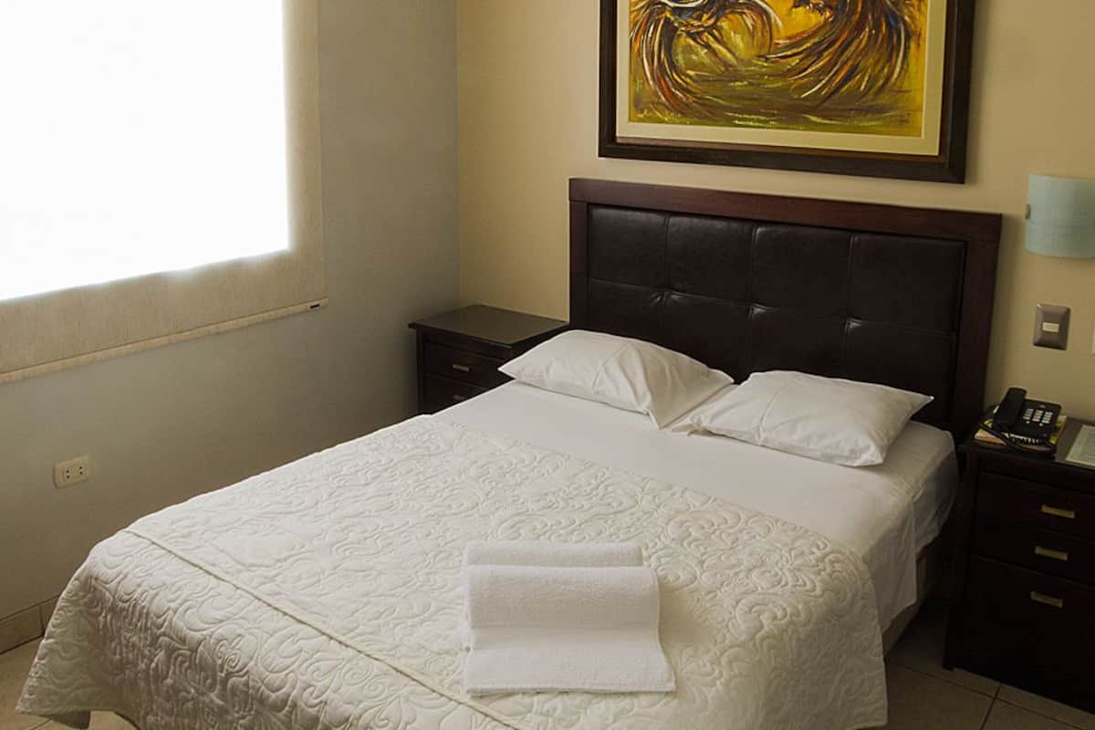 A neatly made bed with a white quilt, a bedside table with a lamp, and a painting on the wall.