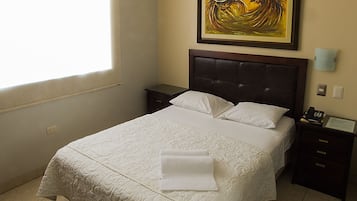 Uma cama bem arrumada com um edredom branco, uma mesa de cabeceira com abajur e um quadro na parede.
