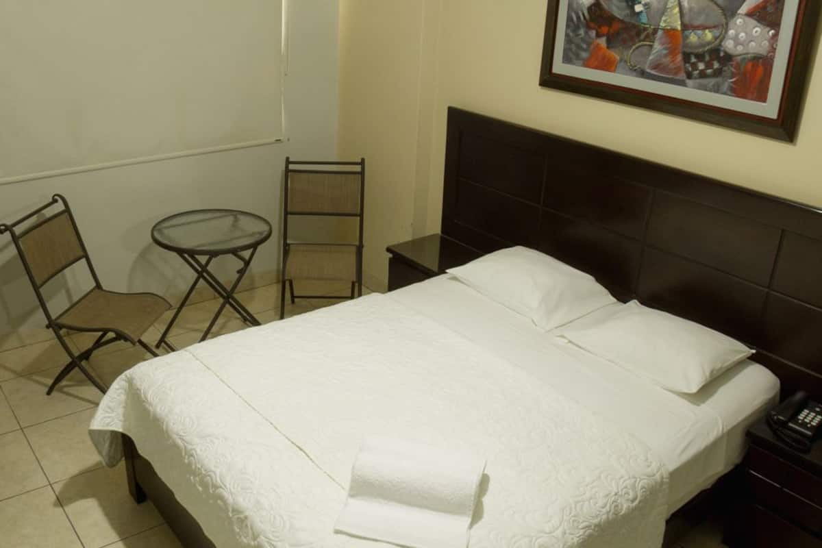 A hotel room with a bed, a small table, a chair, a desk, and a nightstand.