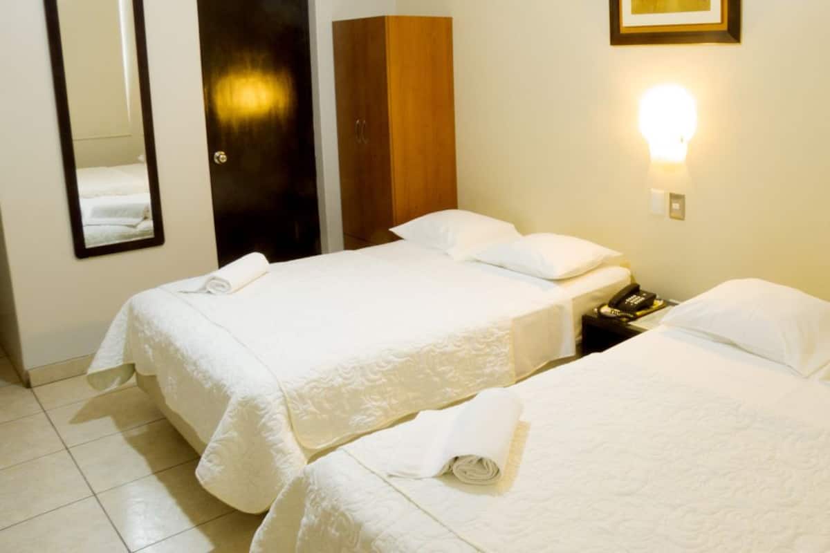 A hotel room with two beds, a wooden wardrobe, a bedside telephone, and a wall-mounted light fixture.