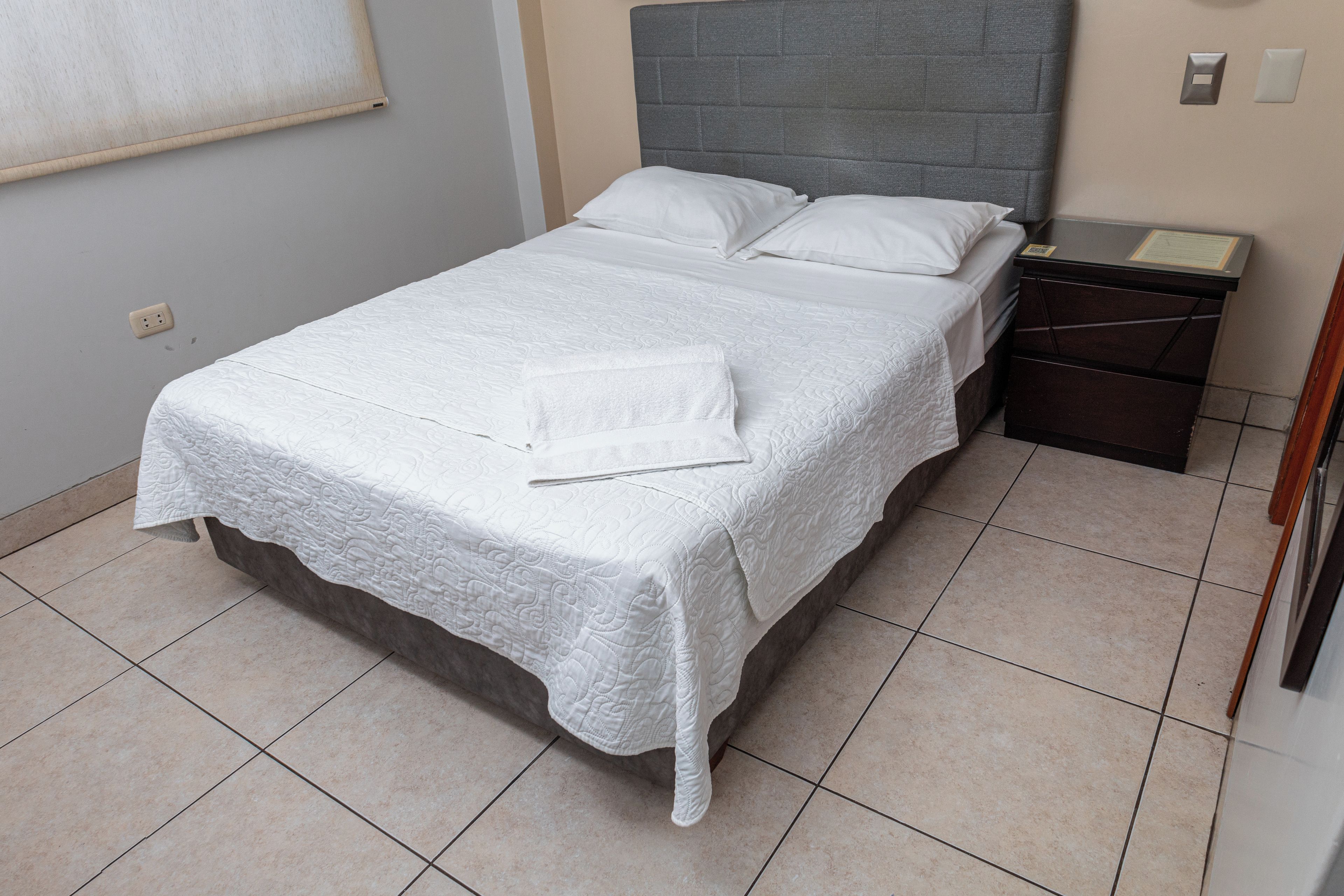 Una cama bien hecha con edredón blanco y almohadas, una mesita de noche con lámpara y un enchufe de pared.