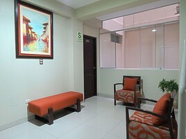 Un área de espera con un banco, dos sillas con cojines naranja, un cuadro enmarcado y una puerta con un letrero.