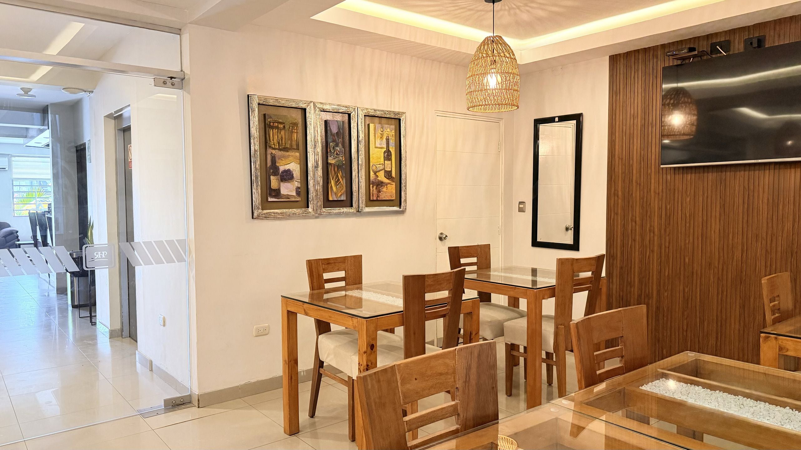 Un comedor con mesa de vidrio y sillas de madera, televisor montado en la pared y cuadros colgados sobre la mesa.