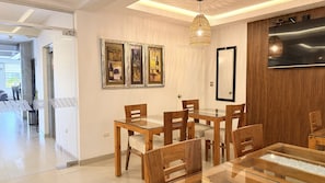 Un comedor con mesa de vidrio y sillas de madera, televisor montado en la pared y cuadros colgados sobre la mesa.