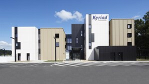 A modern hotel with a prominent 'Kyriad' sign, featuring a mix of white and beige buildings, a central entrance, and a parking lot.