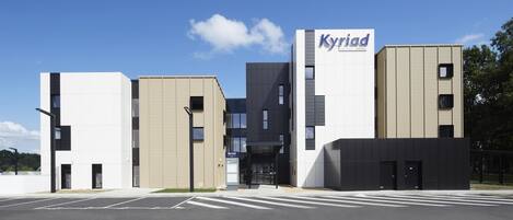 Un hotel moderno con un llamativo letrero de 'Kyriad', con edificios de tonos blanco y beige, una entrada central y un estacionamiento.