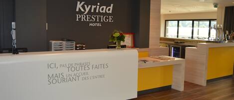 A reception area at the Kyriad Prestige Hotel with a modern design, featuring a reception desk, a framed welcome sign, and a window providing a view of the outdoors.