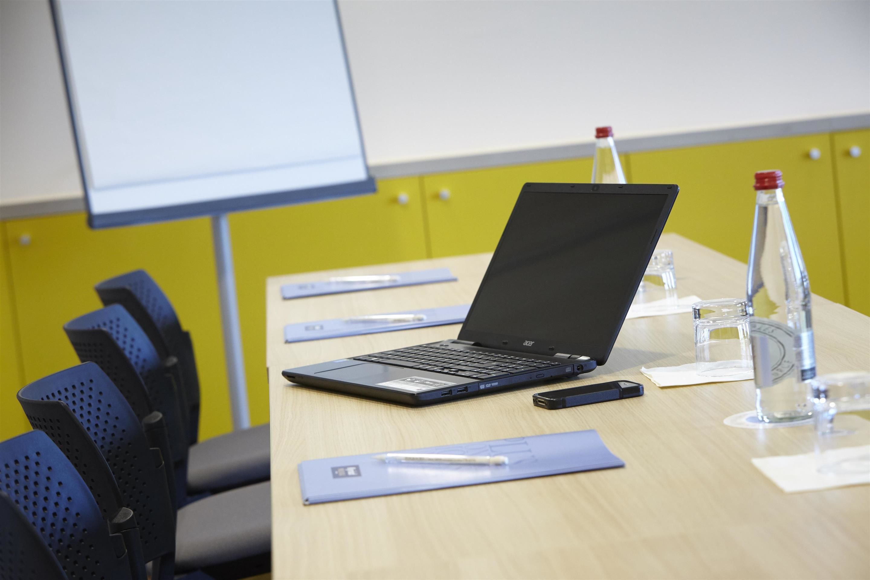 Un salón de conferencias con una laptop sobre una mesa, una pantalla de proyector y una pared amarilla.