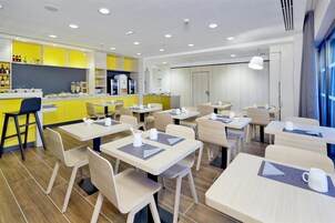 Una cafetería moderna con decoración amarilla y blanca, varias mesas y sillas, y una barra con bebidas y aperitivos.