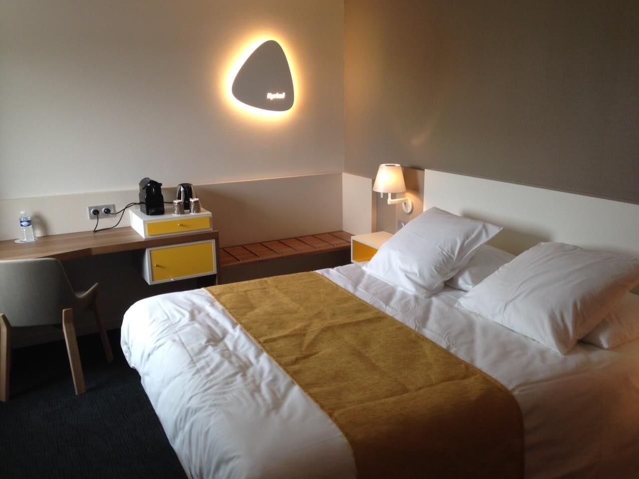 A hotel room with a bed, a desk, a chair, a nightstand, and a wall-mounted lamp.