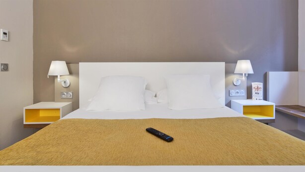 A hotel room with a large bed, two bedside tables, two wall-mounted lamps, and a remote control on the bed.