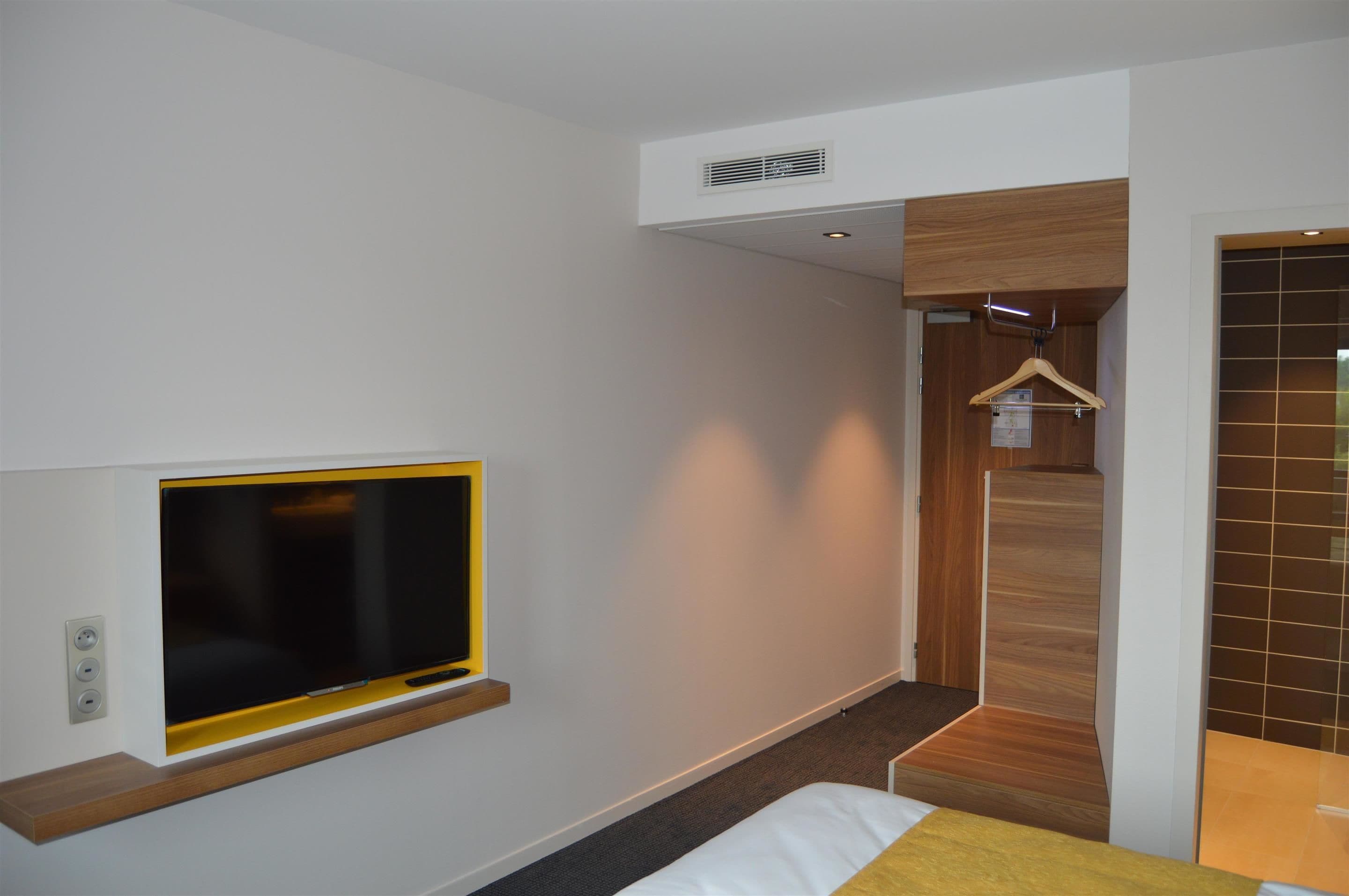 A modern hotel room with a flat-screen TV mounted on the wall, a wooden coat rack, and a small seating area with a bench.