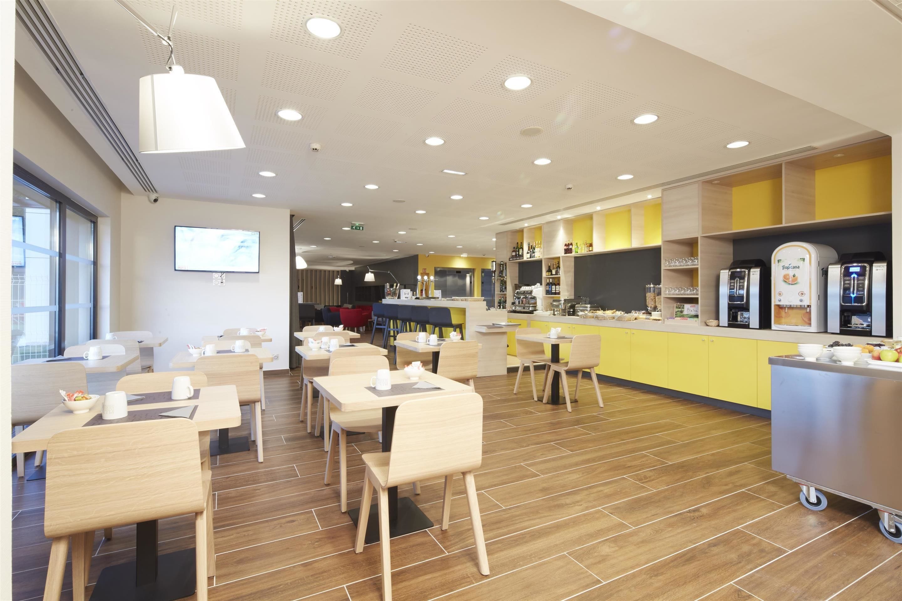 A modern dining area with wooden tables and chairs, a kitchen counter with appliances, and a bright ceiling with recessed lighting.