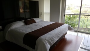 A large bed with a brown throw blanket, a flat-screen TV mounted on the wall, and a balcony with a view of greenery.