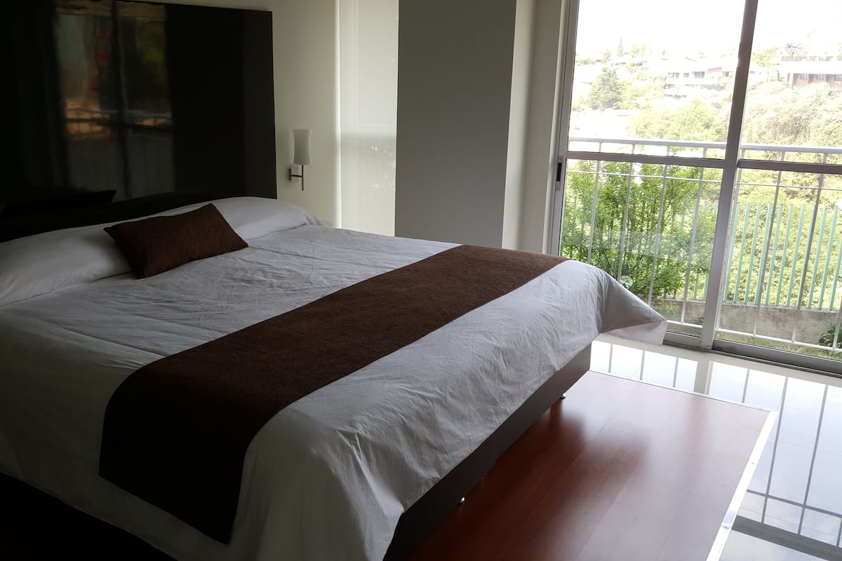 A large bed with a brown throw blanket, a flat-screen TV mounted on the wall, and a balcony with a view of greenery.